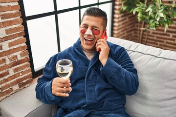 Hispanic young man wearing bathrobe and eye bags patches drinking wine speaking on the phone...