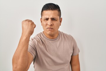 Hispanic young man standing over white background angry and mad raising fist frustrated and furious while shouting with anger. rage and aggressive concept.
