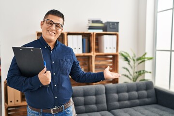 Young latin man business worker holding clipboard doing welcome gesture at office