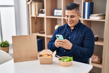 Hispanic young man eating take away food using smartphone smiling with a happy and cool smile on face. showing teeth.