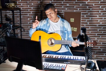 Hispanic young man playing classic guitar at music studio pointing up looking sad and upset, indicating direction with fingers, unhappy and depressed.