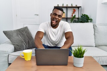 Fototapeta na wymiar Young african man using laptop at home sticking tongue out happy with funny expression. emotion concept.