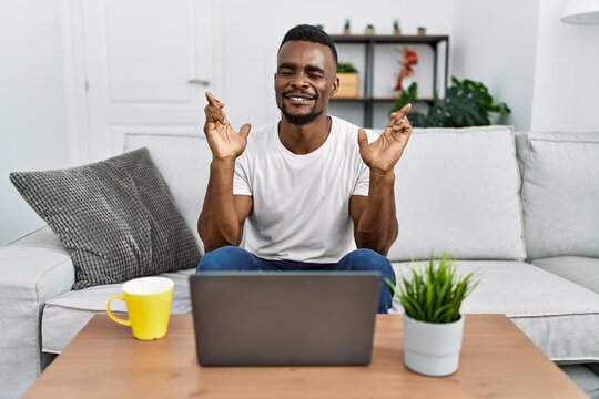 Young african man using laptop at home gesturing finger crossed smiling with hope and eyes closed. luck and superstitious concept.