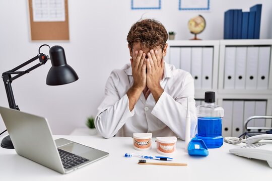 Young hispanic dentist man working at medical clinic with sad expression covering face with hands while crying. depression concept.