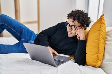 Young hispanic man using laptop lying on bed at bedroom