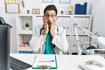 Young man with beard wearing doctor uniform and stethoscope at the clinic afraid and shocked, surprise and amazed expression with hands on face