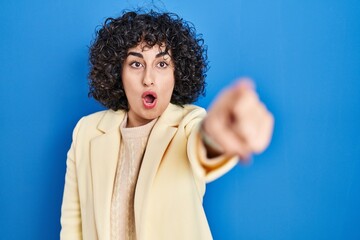 Fototapeta na wymiar Young brunette woman with curly hair standing over blue background pointing with finger surprised ahead, open mouth amazed expression, something on the front