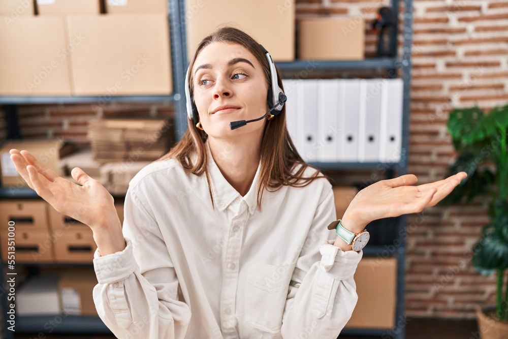 Sticker Young caucasian woman working at small business ecommerce wearing headset clueless and confused expression with arms and hands raised. doubt concept.