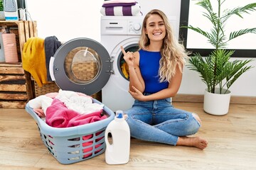 Young beautiful woman doing laundry sitting by wicker basket with a big smile on face, pointing...