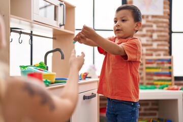 Adorable hispanic toddler playing with play kitchen standing at kindergarten