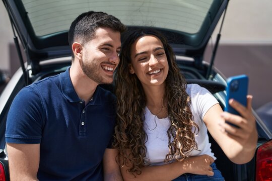 Young hispanic couple sitting on car trunk make selfie by smartphone at street