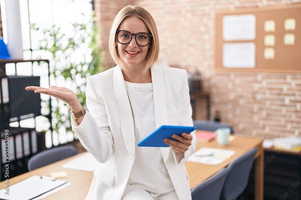 Wall mural Young caucasian woman working at the office wearing glasses smiling cheerful presenting and pointing with palm of hand looking at the camera.