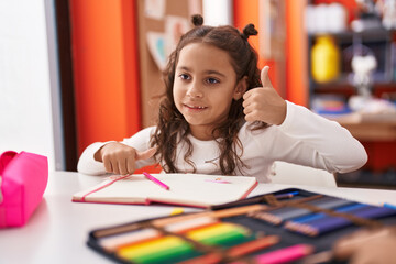 Adorable hispanic girl student sitting on table drawing on notebook at classroom