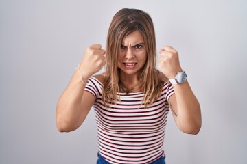 Young hispanic woman standing over isolated background angry and mad raising fists frustrated and...
