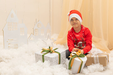 A child in a red Christmas Santa hat sits near New Year's gifts. Happy child in a red New Year's hat plays on a background of Christmas decorations