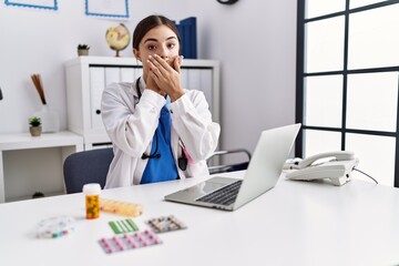 Young hispanic doctor woman wearing doctor uniform working at the clinic shocked covering mouth with hands for mistake. secret concept.