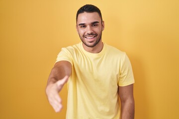 Young hispanic man standing over yellow background smiling friendly offering handshake as greeting and welcoming. successful business.