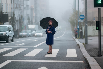 A woman in an overcoat with an umbrella stands by the road in the fog.