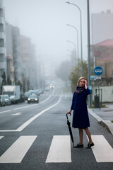 A woman with an umbrella at a crosswalk in overcast weather. - 551354430