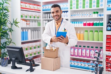 Young hispanic man pharmacist using touchpad holding pills bottle at pharmacy