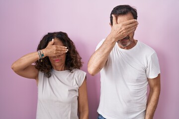 Middle age hispanic couple together over pink background covering eyes with hand, looking serious and sad. sightless, hiding and rejection concept
