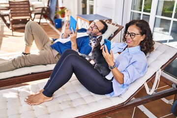 Middle age hispanic couple using smartphone and touchpad lying on hammock with dog at terrace