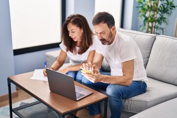 Obraz premium Middle age man and woman couple accounting using smartphone and laptop at home