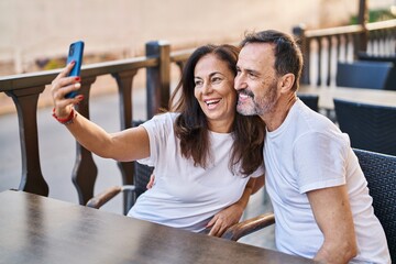 Middle age man and woman couple make selfie by the smartphone sitting on table at coffee shop terrace