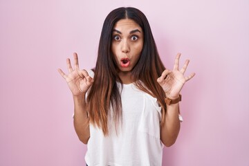 Young arab woman standing over pink background looking surprised and shocked doing ok approval symbol with fingers. crazy expression