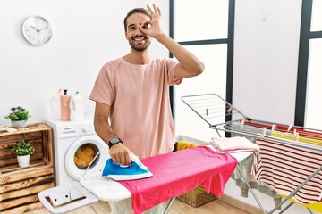 Obraz premium Young hispanic man ironing clothes at home doing ok gesture with hand smiling, eye looking through fingers with happy face.