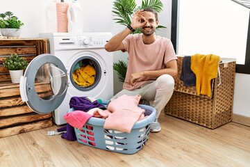 Young hispanic man putting dirty laundry into washing machine smiling happy doing ok sign with hand on eye looking through fingers