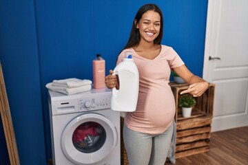Young pregnant woman doing laundry holding detergent bottle pointing finger to one self smiling happy and proud