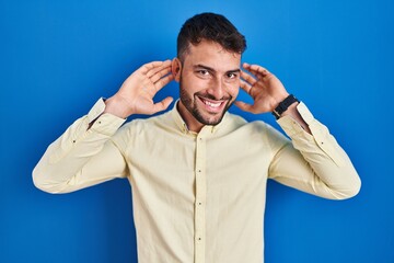 Handsome hispanic man standing over blue background smiling pulling ears with fingers, funny...
