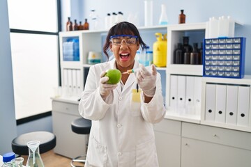 Hispanic woman working at scientist laboratory with apple angry and mad screaming frustrated and furious, shouting with anger. rage and aggressive concept.