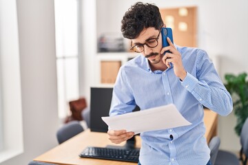 Obraz premium Young caucasian man business worker talking on smartphone reading document at office