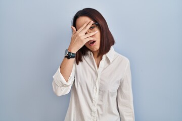 Young hispanic woman standing over white background peeking in shock covering face and eyes with hand, looking through fingers with embarrassed expression.