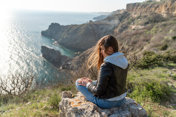 Naklejka na ściany i meble Woman tourist enjoying the sunset over the sea mountain landscape. Sits outdoors on a rock above the sea. She is wearing jeans, a blue hoodie and a black leather jacket. Healthy lifestyle, harmony and