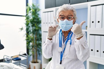 Fototapeta premium Senior woman with grey hair wearing doctor uniform and medical mask holding syringe smiling happy and positive, thumb up doing excellent and approval sign