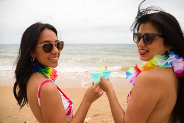 Two young and beautiful latin women with a glass of blue cocktail and toast and celebrate the event with happiness. The women are on the beach with sunglasses. Holiday and travel concept.