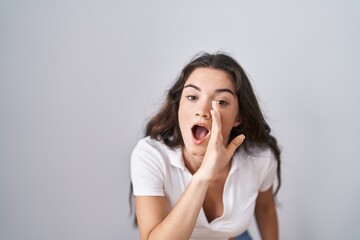 Young teenager girl standing over white background hand on mouth telling secret rumor, whispering malicious talk conversation