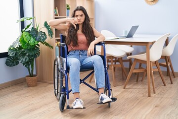 Young teenager girl sitting on wheelchair at the living room with angry face, negative sign showing dislike with thumbs down, rejection concept