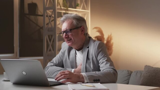 Bad work report. Stressed aged man. Heart pain. Worried desperate male closing laptop holding hand on chest feeling attack in light shadow room interior.