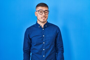 Young hispanic man wearing glasses over blue background winking looking at the camera with sexy expression, cheerful and happy face.