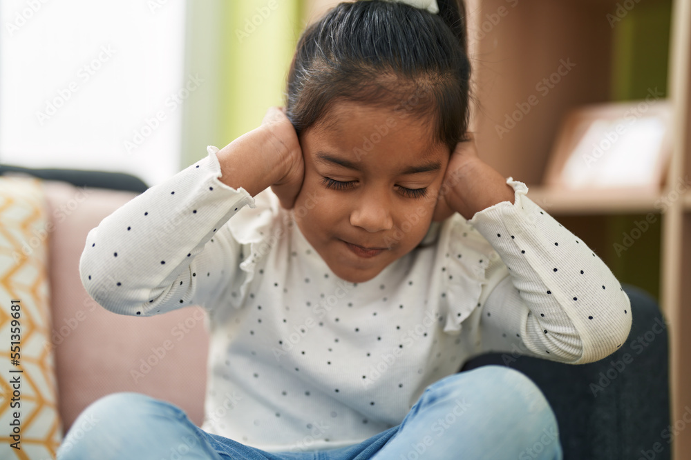 Sticker Adorable hispanic girl sitting on sofa with hands on ears at home