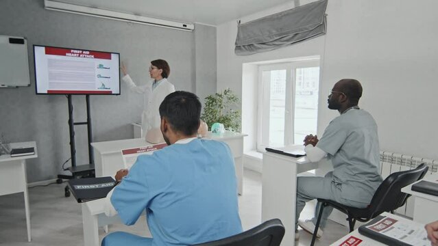 Mid adult paramedic specialist pointing at screen speaking about first aid rules at medical training, ethnically diverse group of doctors and nurses carefully listening to her