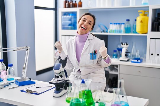 Young brunette woman working at scientist laboratory very happy and excited doing winner gesture with arms raised, smiling and screaming for success. celebration concept.
