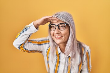 Middle age woman with grey hair standing over yellow background wearing glasses very happy and...