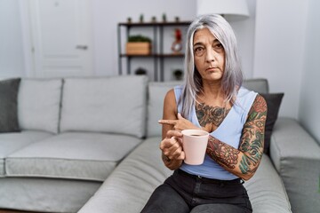 Middle age grey-haired woman drinking coffee sitting on the sofa at home pointing with hand finger to the side showing advertisement, serious and calm face