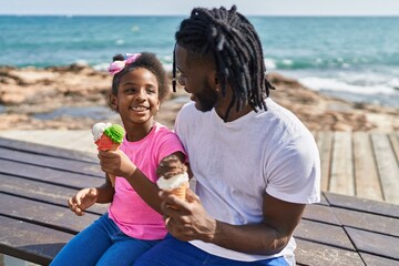 Father and daughter eating ice cream sitting together on bench at seaside
