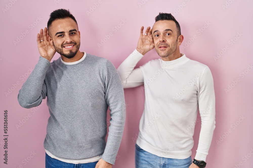 Poster homosexual couple standing over pink background smiling with hand over ear listening an hearing to r